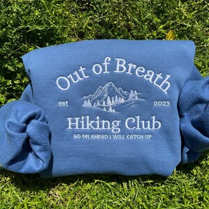 May include: A blue sweatshirt featuring white embroidery that reads Out of Breath Hiking Club, est 2023, and Go on ahead I will catch up, with a central graphic of a mountain range and pine trees. The design is centered on the chest area.
