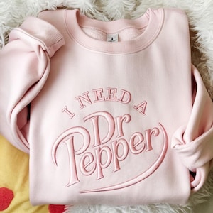Puede incluir: Una sudadera rosa claro con la frase bordada "I NEED A Dr Pepper" en un color rosa a juego. La sudadera tiene cuello redondo y mangas largas.