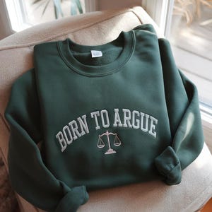 May include: A dark green sweatshirt with the text "BORN TO ARGUE" in white, arched across the chest. Below the text is a small white scale graphic. The sweatshirt is folded on a beige armchair.