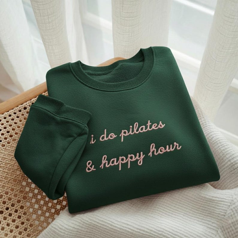 May include: A dark green sweatshirt with the embroidered text "i do pilates & happy hour" in a light pink script. The sweatshirt is folded on a woven chair with a white blanket.