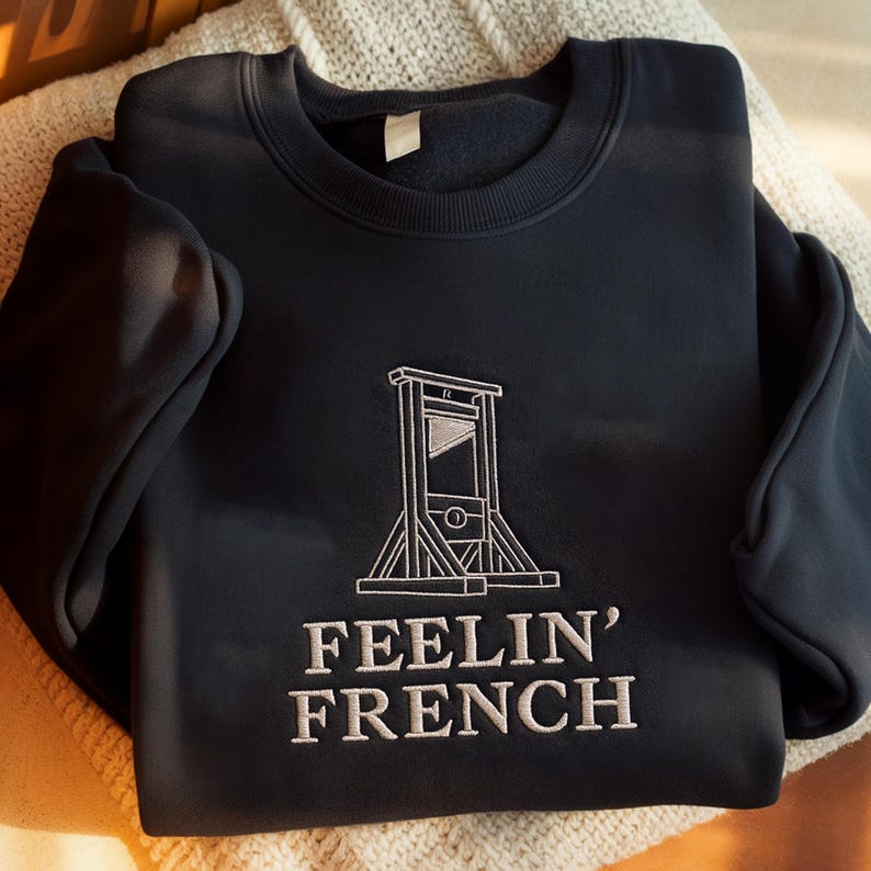 May include: A dark navy blue crewneck sweatshirt featuring white embroidery on the chest. The design depicts a guillotine illustration positioned above the text "FEELIN' FRENCH" in a bold, capitalized, serif font style.