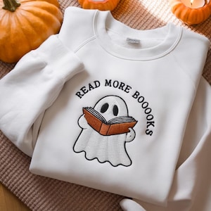 Peut inclure: Sweat-shirt blanc avec l'inscription brodée "READ MORE BOOOOKS" autour d'un fantôme de dessin animé lisant un livre. Le fantôme est blanc avec des yeux noirs et tient un livre orange. Le sweat-shirt a un col rond et des manches longues.
