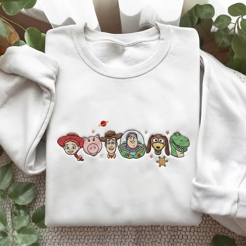 May include: A white crewneck sweatshirt featuring an embroidered design of six animated characters from Toy Story, including a cowgirl, a pig, a cowboy, a space ranger, a dachshund, and a dinosaur, accented with small stars and a planet.