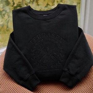 May include: Black crewneck sweatshirt with the word "TERRASEN" embroidered across the top. Below the text is a circular design with a mountain scene and the words "HOME OF THE STAGS". The sweatshirt is folded on a pink surface.