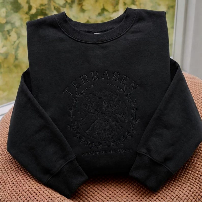May include: Black crewneck sweatshirt with the word "TERRASEN" embroidered across the top. Below the text is a circular design with a mountain scene and the words "HOME OF THE STAGS". The sweatshirt is folded on a pink surface.