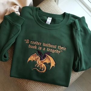 May include: A dark green sweatshirt with embroidered text and a dragon design. The text reads, "A reader without their book is a tragedy." The dragon is gold and black with outstretched wings. The sweatshirt is folded on a neutral surface.