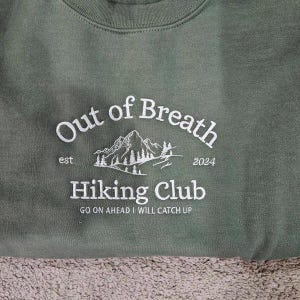 May include: An olive green sweatshirt featuring white embroidered text that reads Out of Breath Hiking Club, est 2024, and Go on ahead I will catch up. The design includes a white line art illustration of a mountain range with pine trees.
