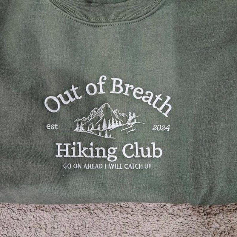 May include: An olive green sweatshirt featuring white embroidered text that reads Out of Breath Hiking Club, est 2024, and Go on ahead I will catch up. The design includes a white line art illustration of a mountain range with pine trees.