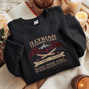 Puede incluir: Sudadera negra con texto y gráficos bordados. El diseño incluye las palabras "ILLYRIAN TRAINING CAMP", una montaña, espadas cruzadas y las frases "NO MERCY. NO LIMITS." y "BLOOD. SWEAT. WINGS."