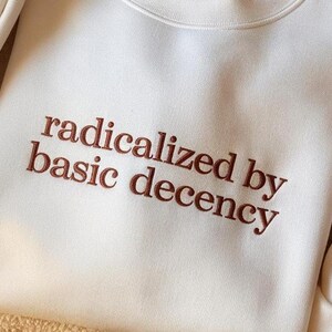 May include: A cream-colored crewneck sweatshirt featuring the embroidered text "radicalized by basic decency" in a dark brown, serif font. The garment has long sleeves and a ribbed neckline, showcasing a clean, simple design with clear lettering.