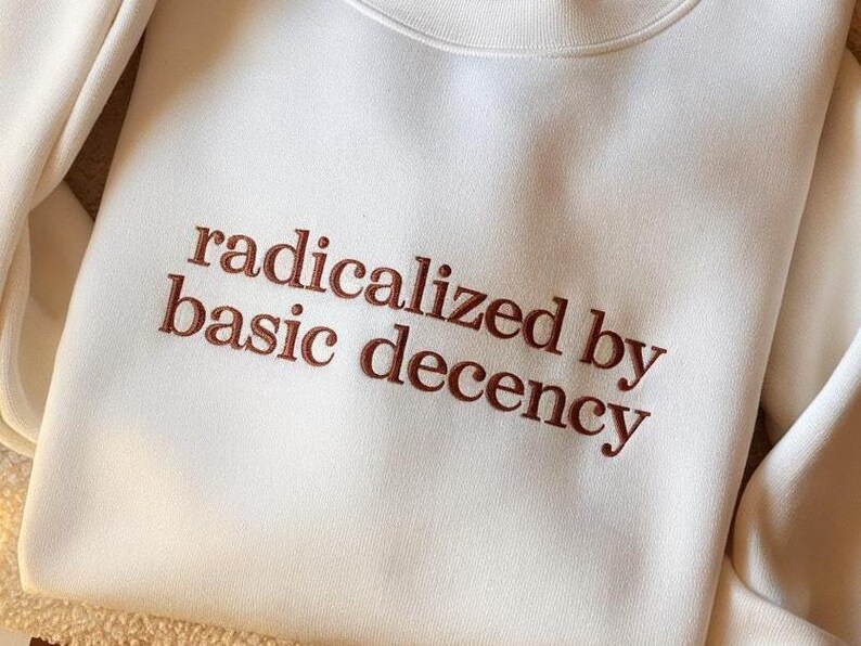 May include: A cream-colored crewneck sweatshirt featuring the embroidered text "radicalized by basic decency" in a dark brown, serif font. The garment has long sleeves and a ribbed neckline, showcasing a clean, simple design with clear lettering.