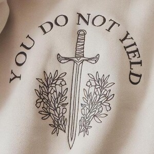 May include: A beige hoodie with a black graphic design featuring a sword with a floral design on either side. The text "You Do Not Yield" is written in a circle around the sword.