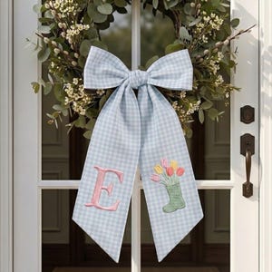 Può includere: Una ghirlanda primaverile con un fiocco a quadretti azzurro e bianco. Il fiocco presenta una lettera "E" ricamata in rosa e uno stivale verde con tulipani ricamati. La ghirlanda è fatta di verde e fiori bianchi, appesa a una porta bianca.