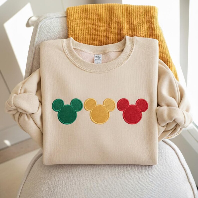 Puede incluir: Una sudadera color crema con tres dise&ntilde;os de cabeza de Mickey Mouse en verde, amarillo y rojo. La sudadera est&aacute; doblada sobre una silla con una manta amarilla sobre el respaldo. El dise&ntilde;o es bordado.