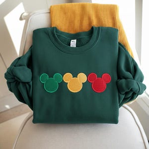 Puede incluir: Una sudadera verde oscuro con tres dise&ntilde;os de cabeza de Mickey Mouse en verde, amarillo y rojo. La sudadera est&aacute; doblada sobre una silla con una bufanda amarilla en el fondo. Las mangas est&aacute;n enrolladas.