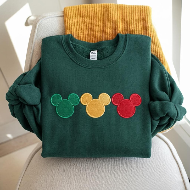 Puede incluir: Una sudadera verde oscuro con tres dise&ntilde;os de cabeza de Mickey Mouse en verde, amarillo y rojo. La sudadera est&aacute; doblada sobre una silla con una bufanda amarilla en el fondo. Las mangas est&aacute;n enrolladas.