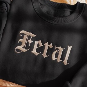May include: Black sweatshirt with the word "Feral" embroidered in a vintage-style font. The text is in a light beige color, contrasting against the dark fabric. The sweatshirt has a crew neck.