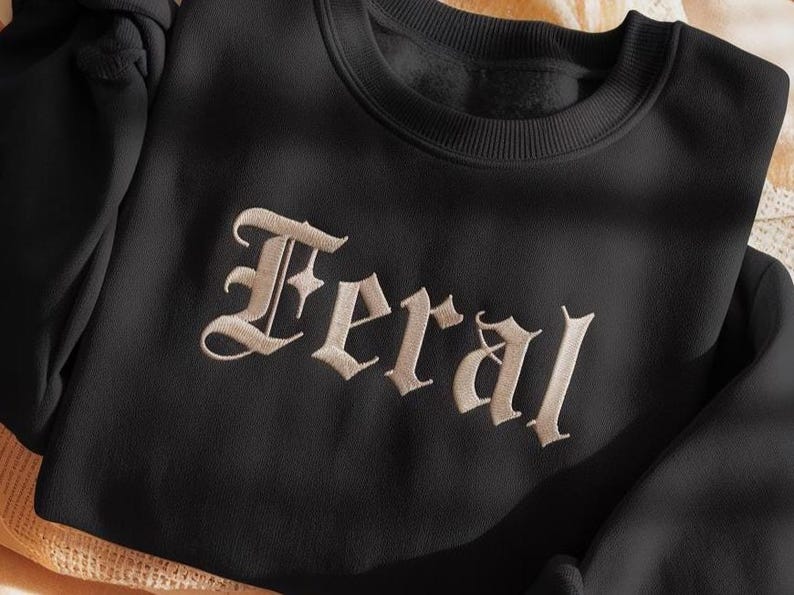 May include: Black sweatshirt with the word "Feral" embroidered in a vintage-style font. The text is in a light beige color, contrasting against the dark fabric. The sweatshirt has a crew neck.