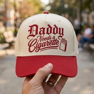 Könnte beinhalten: Eine cremefarbene Baseballkappe mit rotem Schirm. Auf der Vorderseite befindet sich ein roter Stickschriftzug mit dem Text Daddy Needs a Cigarette, ergänzt durch eine kleine rote Grafik einer Zigarettenschachtel. Die Kappe hat eine feste Krone.