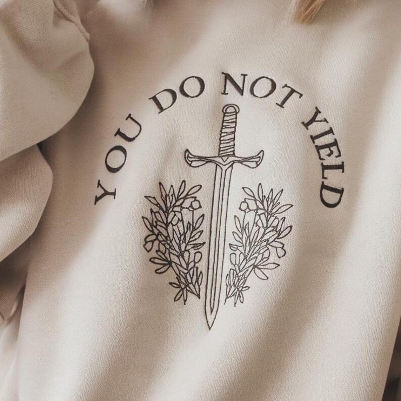 May include: A beige sweatshirt with a black graphic design of a sword with leaves and the text "You Do Not Yield".