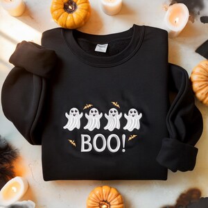 May include: A black crewneck sweatshirt featuring an embroidered design of four white ghosts with smiling faces and small orange bats. Below the ghosts, the text BOO! is embroidered in white. The sweatshirt has long sleeves and a ribbed collar.