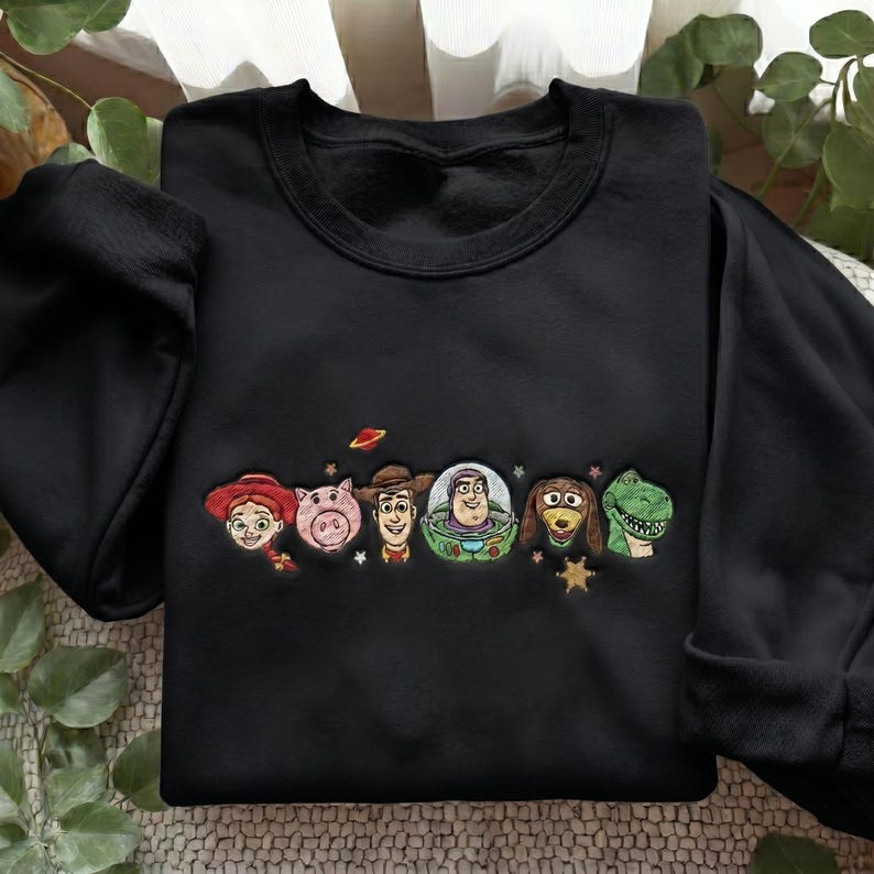 May include: A black crewneck sweatshirt featuring an embroidered design of six animated characters including a cowgirl, a pig, a cowboy, a space ranger, a dachshund, and a green dinosaur, accented with small embroidered stars and a planet.