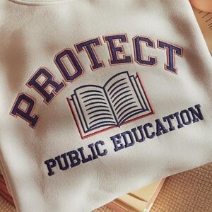 May include: A beige sweatshirt featuring dark blue embroidered text that reads PROTECT PUBLIC EDUCATION in an arched layout. Centered between the two lines of text is an embroidered graphic of an open book with dark blue outlines.