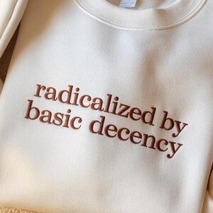 May include: A cream-colored crewneck sweatshirt featuring the embroidered text "radicalized by basic decency" in a dark brown, serif font. The garment has a ribbed collar and a smooth, solid-colored fabric construction with visible stitching.