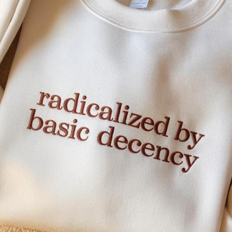 May include: A cream-colored crewneck sweatshirt featuring the embroidered text "radicalized by basic decency" in a dark brown, serif font. The garment has a ribbed collar and a smooth, solid-colored fabric construction with visible stitching.