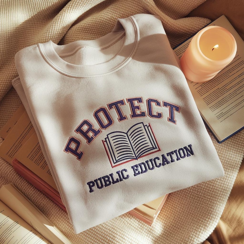 May include: A cream-coloured crewneck sweatshirt featuring an embroidered design with the text "PROTECT PUBLIC EDUCATION" in dark blue letters arched over a graphic of an open book with red and blue outlines, centred on the chest of the garment.