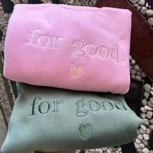 以下が含まれることがあります： ピンクとグリーンの2つのスウェットシャツ。前面に「for good.」という文字が刺繍されています。両方のスウェットシャツには、テキストの下に小さなハートが刺繍されています。