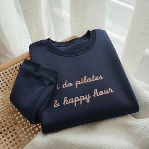 May include: Navy blue sweatshirt with the text "i do pilates & happy hour" in a cursive font. The long-sleeved top is folded on a woven chair. The fabric appears soft and comfortable.