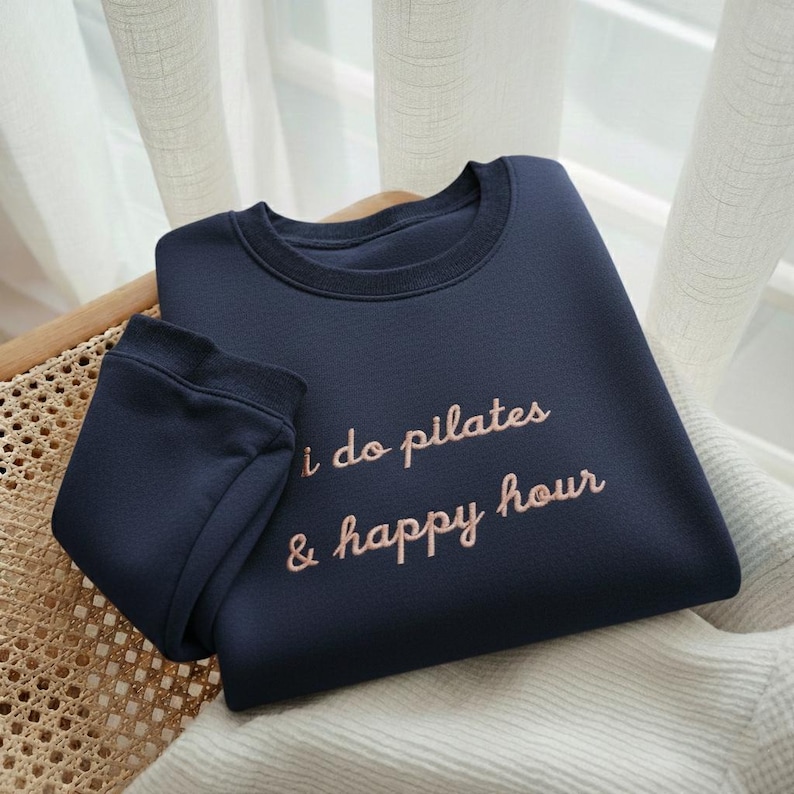 May include: Navy blue sweatshirt with the text "i do pilates & happy hour" in a cursive font. The long-sleeved top is folded on a woven chair. The fabric appears soft and comfortable.
