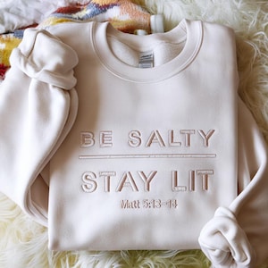 以下が含まれることがあります： BE SALTY STAY LITおよびMatt 5:13-14という刺繍テキストが施されたクリーム色のクルーネックトレーナー。刺繍は同系色のクリーム色の糸で仕上げられており、長袖の衣類の前面に繊細な同系色の効果を生み出しています。