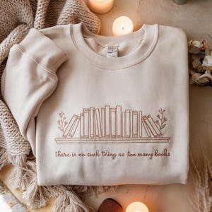 May include: A beige sweatshirt with a brown embroidered design of a bookshelf overflowing with books. The text "there is no such thing as too many books" is embroidered below the bookshelf.