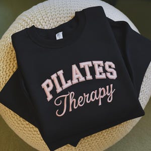 May include: Black crewneck sweatshirt with the words "PILATES Therapy" embroidered in a light pink, arched font. The sweatshirt is folded neatly on a textured, off-white surface. The overall aesthetic is clean and modern.