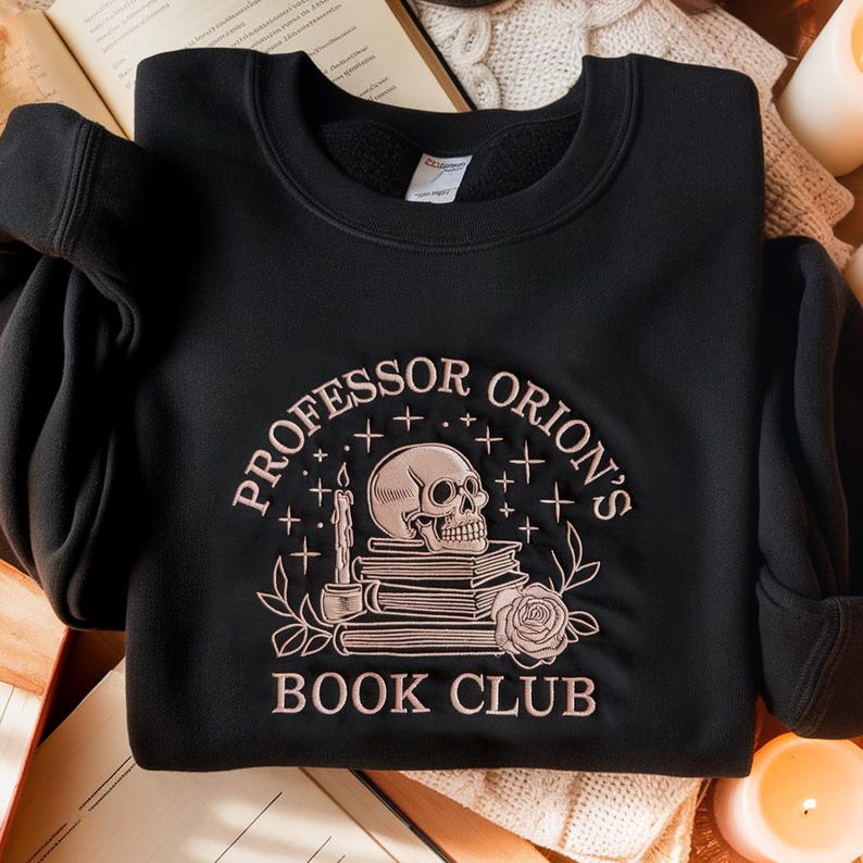 May include: Black sweatshirt with the embroidered text "Professor Orion's Book Club." The design features a skull, books, a candle, and a rose in a pinkish-beige color. The sweatshirt is made of a soft material.