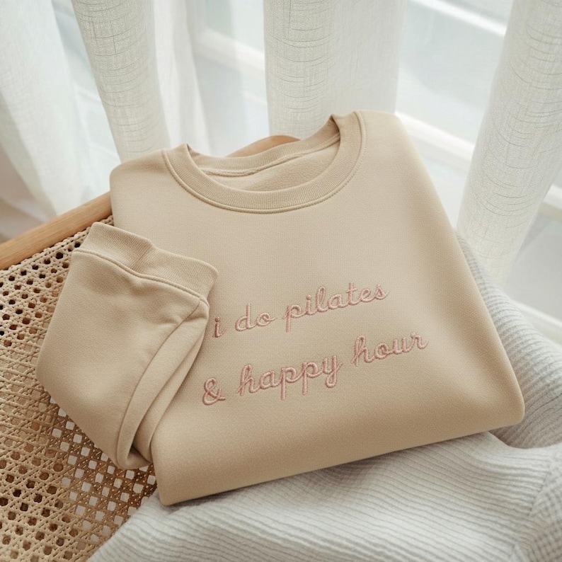 May include: Beige sweatshirt with the embroidered text "i do pilates & happy hour" in a light pink color. The sweatshirt is folded and placed on a wicker chair. The sleeves are folded over.