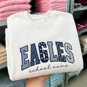 Peut inclure: Un sweat-shirt à col rond couleur crème avec le mot EAGLES en grandes lettres style bloc. Chaque lettre présente un motif bleu foncé unique, incluant des rayures, des pois, des étoiles, du vichy et des fleurs. Les mots school name sont en dessous.