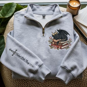 May include: A light gray quarter-zip sweatshirt featuring an embroidered black dragon with a gold crown resting on a stack of books surrounded by flowers. The right sleeve displays an embroidered sword with the text Violent Little Thing.