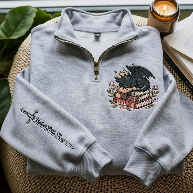 May include: A light gray quarter-zip sweatshirt featuring an embroidered black dragon with a gold crown resting on a stack of books surrounded by flowers. The right sleeve displays an embroidered sword with the text Violent Little Thing.