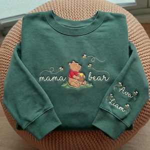 Könnte beinhalten: Ein dunkelgrünes Sweatshirt mit gesticktem Winnie-Puuh-Design. Das Design enthält die Wörter "Mama Bear" und "Ava, Liam" mit Bienen. Winnie Puuh hält einen Honigtopf.