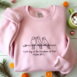 May include: A light pink sweatshirt with a crew neck and long sleeves. The front features a black line drawing of three birds perched on a branch, with the text "I will sing of the Goodness of God -Psalm 89:1."