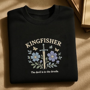 Può includere: Felpa nera con un designo ricamato bianco che presenta una spada, fiori e il testo "KINGFISHER". Il testo "The devil is in the details." è anche ricamato sulla felpa.