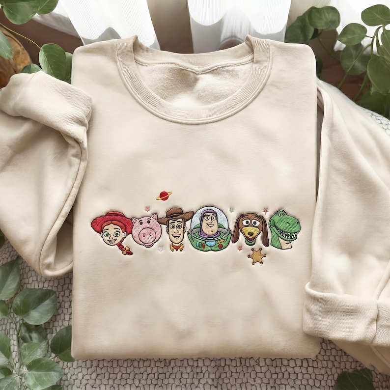 May include: A beige crewneck sweatshirt featuring an embroidered design of six animated characters from Toy Story, including a cowgirl, a pig, a cowboy, a space ranger, a dachshund, and a green dinosaur, accented with small stars and a planet.
