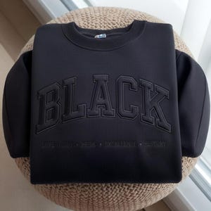 May include: Black sweatshirt with the word "BLACK" in large, raised lettering across the chest. Below the word "BLACK" are the words "LOVE JOY PRIDE EXCELLENCE HISTORY" in smaller lettering. The sweatshirt is folded on a woven surface.