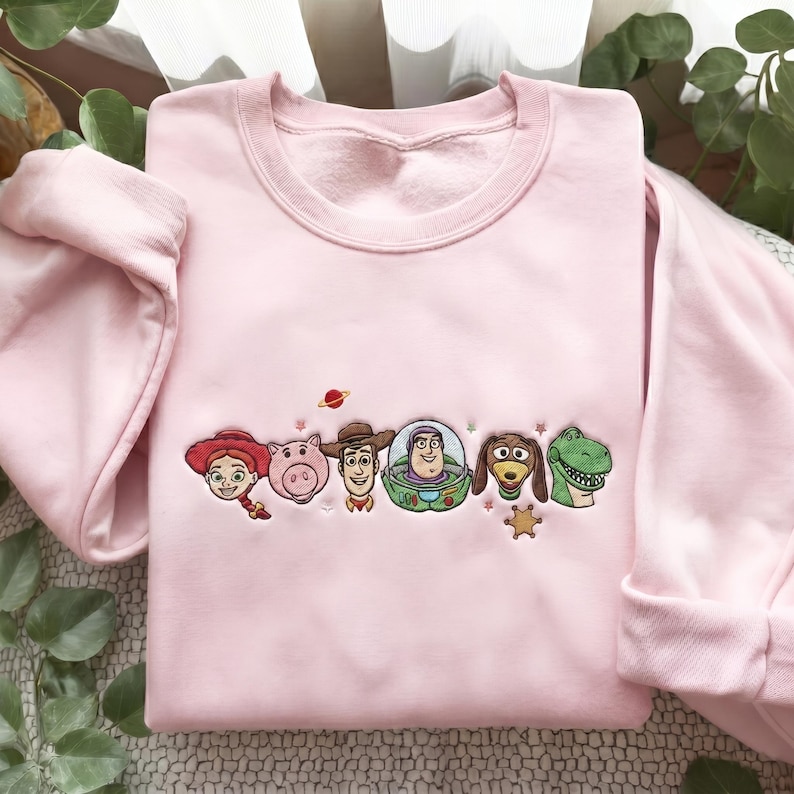 May include: A light pink crewneck sweatshirt featuring an embroidered design of six animated characters including a cowgirl, a pig, a cowboy, a space ranger, a dachshund, and a green dinosaur, accented with small stars and a planet.