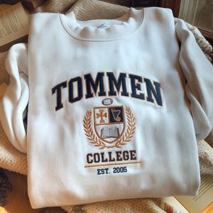 May include: A white crewneck sweatshirt with the word "TOMMEN" in navy blue arched across the chest. Below the word is a college crest with the word "COLLEGE" and the year "EST. 2005" in navy blue.