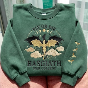 Peut inclure: Sweat-shirt vert avec un motif de dragon brodé. Le motif comprend le texte "FLY OR DIE" au-dessus d'un dragon aux ailes dorées, et "BASGIATH WAR COLLEGE" en dessous. Des silhouettes de dragons dorés ornent la manche.