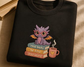 Könnte beinhalten: Ein schwarzes Sweatshirt mit Rundhalsausschnitt und einer Stickerei eines violetten Drachen mit rosa Brille, der auf drei Büchern mit der Aufschrift read books, be kind, stay weird sitzt, neben einer rosa Tasse mit gelbem Herz.